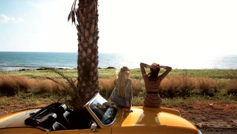 Young-women-sitting-on-retro-convertible-car-looking-at-sea