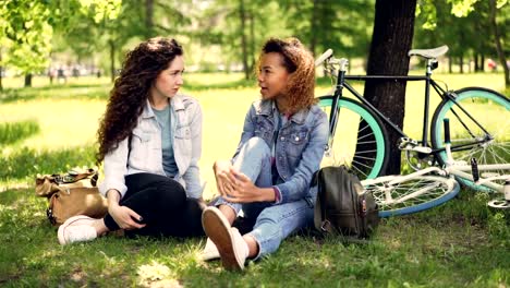 African-American-girl-is-talking-to-her-Caucasian-friend-sitting-on-lawn-in-park,-modern-bikes-and-backpacks-are-visible.-Mixed-race-friendship-and-conversation-concept.