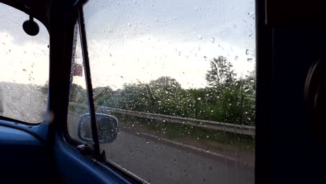 View-through-the-side-window-of-the-old-retro-car-while-moving-at-rainy-weather.-Raindrops-falling-on-the-glass-of-vintage-automobile-during-drive-at-countryside.-Close-up-Slow-motion