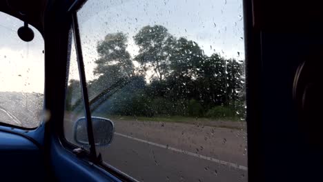 View-through-the-side-window-of-the-old-retro-car-while-moving-at-rainy-weather.-Raindrops-falling-on-the-glass-of-vintage-automobile-during-drive-at-countryside.-Close-up-Slow-motion