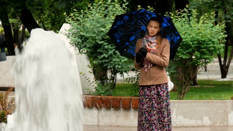 Una-hermosa-chica-con-ojos-tristes-está-de-pie-bajo-la-lluvia-en-el-parque-de-la-ciudad,-cerca-de-la-fuente.