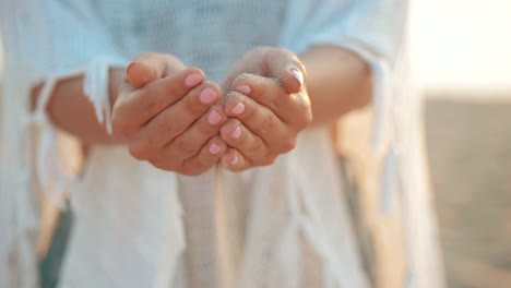 Close-up-of-woman-pouring-sand-running-through-fingers-at-the-beach.-Hand-of-a-woman-in-white-clothing-drizzling-sea-sand-through-her-fingers-against-an-ocean-backdrop,-conceptual-image