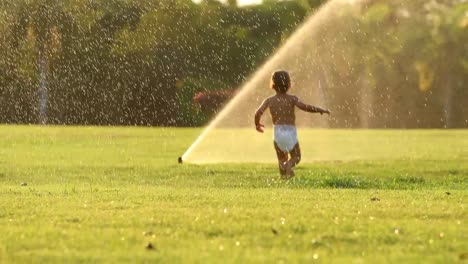 Dreamy-idealic-scene-of-infant-toddler-boy-running-in-green-field-with-water-sprinkles-during-sunset-golden-hour-time-in-4k-clip-resolution