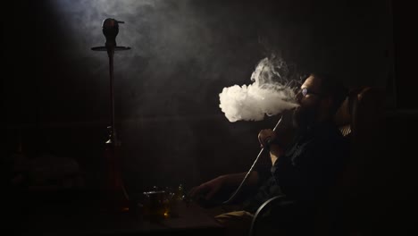 young-bearded-man-in-glasses-smokes-a-hookah-and-blow-out-smoke-closeup-on-black-background-in-slow-motion-in-4k