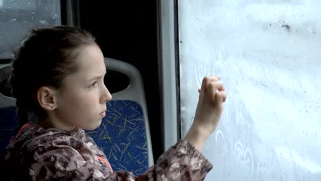 Ein-kleines-Mädchen-sitzt-im-Bus-auf-dem-Rücksitz-und-zeichnet-Bilder-mit-dem-Finger-auf-dem-nassen-beschlagene-Glas.-Außerhalb-der-Fenster-Auto-fahren.-Porträt.