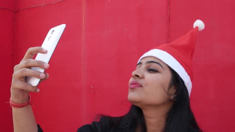 Indian-woman-with-Santa's-hat-taking-selfies-photo-with--mobile-cell-phone,-cheerful-and-excited,-with-red-background