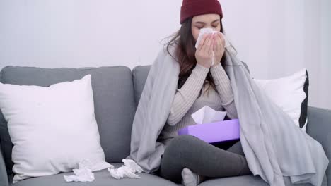 Mujer-asiática-joven-enferma-siente-dolor-de-cabeza-sentado-en-una-silla-envuelta-en-una-manta-gris-en-su-sala-de-estar-en-casa.-Mujer-hermosa-en-el-festival-invierno-y-Navidad-soplar-la-nariz-y-utilice-tejido.