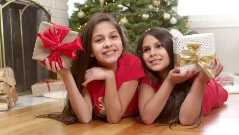 Two-sisters-lay-on-the-floor-in-front-of-the-christmas-tree-and-hold-up-their-presents.-Portrait