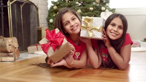 Two-sisters-lay-on-the-floor-in-front-of-the-christmas-tree-and-shake-their-presents.-Portrait