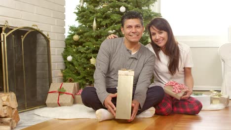 A-couple,-maybe-a-mother-and-father-sit-under-their-christmas-tree-holding-presents-and-smile-into-the-camera