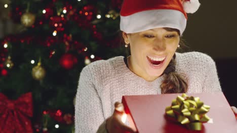 Mädchen-mit-weihnachtsmütze-macht-Wünsche-und-öffnet-ein-Weihnachts-Geschenk-Paket.-Konzept-der-Feiertage-und-Neujahr.-Das-Mädchen-freut-sich-und-lächelt-mit-Weihnachts-Geschenk-in-der-hand.