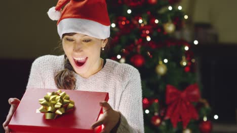 Muchacha-con-sombrero-de-la-Navidad-hace-deseos-y-abre-un-paquete-de-regalo-de-Navidad.-concepto-de-vacaciones-y-año-nuevo.-la-niña-es-feliz-y-sonríe-de-la-mano-con-el-regalo-de-Navidad.