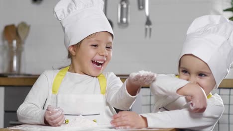 Dos-niñas-en-la-cocina-preparan-comida,-un-postre-para-la-familia.-Como-aprender-a-cocinar-empiezan-jugando-con-harina-y-sonriendo-unos-a-otros.