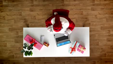 Santa-Claus-working-at-office-on-laptop,-listening-music-in-headphones,-typing,-aerial-view,-top-down-shot,-writes-text-to-a-notebook