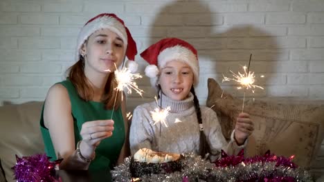 Glückliche-Familie,-zwei-Mädchen,-kleine-und-Teenager-in-den-Hut-des-Weihnachtsmannes-mit-brennenden-Wunderkerzen-auf-den-Kuchen-auf-dem-Sofa-sitzen,-sieht-ihn-an-und-lächelte.-Weihnachten.