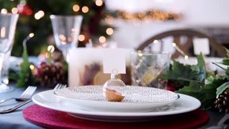 Close-up-panning-shot-of-a-bauble-name-card-holder-arranged-on-a-plate-on-a-Christmas-dining-table,-bokeh-Christmas-lights-in-the-background