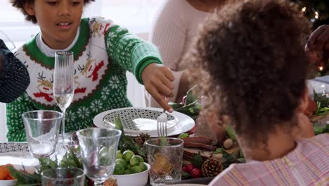 Multi-generation-mixed-race-family-sitting-at-Christmas-dinner-table-eating-together,-panning-shot