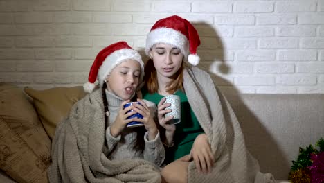 Children,-two-girls,-covered-with-a-blanket,-singing-sitting-in-Santa's-hats-on-the-couch-against-a-white-wall,-in-the-flickering-light.-Portrait.-4K.-25-fps.