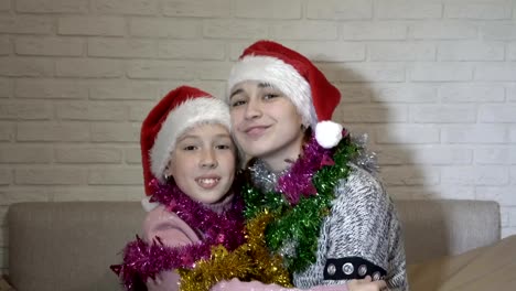 Portrait-of-two-happy-children-girls-in-Santa-Claus-hats-with-Christmas-tinsel-on-their-shoulders,-they-sit-on-the-couch,-hug,-look-at-the-camera-and-smile.-Close-up.