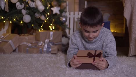 A-little-boy-opens-a-box-with-a-gift-and-rejoices-lying-on-the-floor-near-the-Christmas-tree.-4K