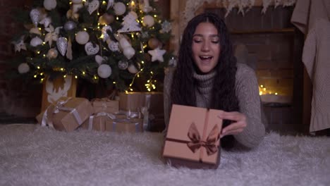 La-chica-abre-la-caja-con-un-regalo-y-regocija-en-el-suelo-cerca-del-árbol-de-Navidad