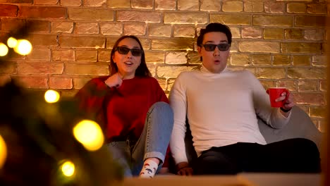 Kaukasische-paar-in-3D-Brille-auf-Sofa-sitzen-und-Film-mit-Tasse-Getränk-wird-in-weihnachtliche-Stimmung-schockiert.