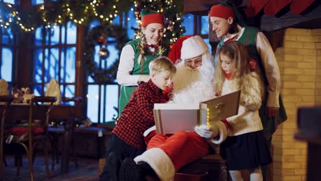 Santa-Claus-está-sentado-en-una-silla-rodeada-por-dos-duendes-y-un-niño-pequeño-con-una-chica-y-teniendo-en-cuenta-un-álbum-de-vacaciones-con-fotos-en-el-fondo-de-adornos-de-año-nuevo-y-el-árbol-de-Navidad-en-la-sala-en-casa