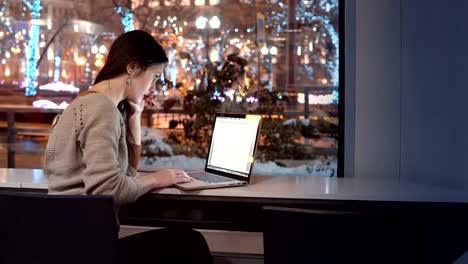 Attractive-young-businesswoman-working-on-laptop-sitting-at-the-bar,-outside-winter-night-city-decorated-for-Christmas