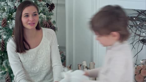 Little-boy-riding-wooden-rocking-horse,-while-happy-mother-watching-him-sitting-near-the-Christmas-tree