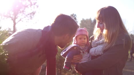 Un-joven-papá-habla-con-su-niña-mientras-mamá-la-sostiene,-en-una-granja-de-árboles-de-Navidad,-la-lente-de-destello