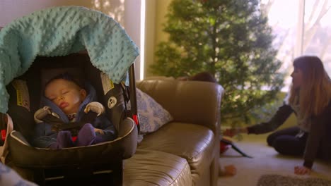 El-bebé-duerme-en-su-asiento-de-coche-en-el-sofá-mientras-que-mamá-y-papá-establecen-el-árbol-de-Navidad