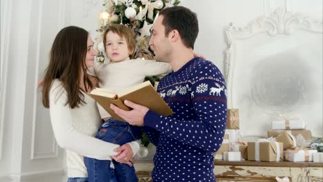 Feliz-padre-leyendo-un-cuento-de-Navidad-mientras-madre-sosteniendo-a-su-alegre-hijo-delante-del-árbol-navideño