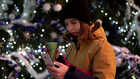 Young-woman-with-cup-of-hot-tea-using-her-smartphone-standing-near-the-christmas-tree.