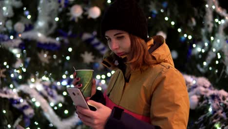 Mujer-joven-con-taza-de-té-caliente-con-su-smartphone-de-pie-cerca-del-árbol-de-Navidad.