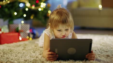 Niedliche-kleine-Mädchen-mit-Tablet-Computer-in-ihren-Händen-liegt-auf-dem-Teppich-unter-dem-Weihnachtsbaum.