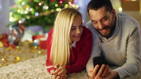 Glückliches-Paar-liegt-auf-dem-Teppich-unter-Weihnachtsbaum,-schauen-Sie-sich-etwas-Interessantes-auf-dem-Smartphone.