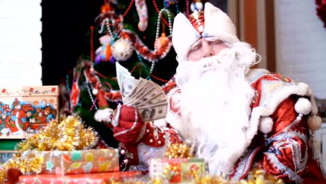 Santa-Claus,-Weihnachtsmann,-Väterchen-Frost-fächelt-sich-mit-einem-Fan-von-Dollar,-er-sitzt-an-einem-Tisch-unter-die-Weihnachtsgeschenke,-im-Hintergrund-ist-ein-Weihnachtsbaum