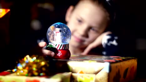 In-the-twilight-of-the-night,-among-the-gifts-in-bright-colorful-paper-packages,-a-pretty-little-blonde-girl-with-a-pink-bow-in-her-hair-and-a-beautiful-dress,-looks-carefully-and-admires-snow-globe.-toy-Christmas-ball-with-sparkles-and-a-snowman-inside