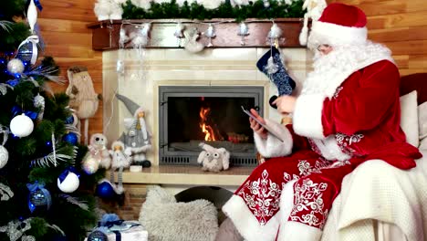 santa-using-tablet,-papa-noel-sitting-in-chair-with-electronic-device,-room-with-fireplace