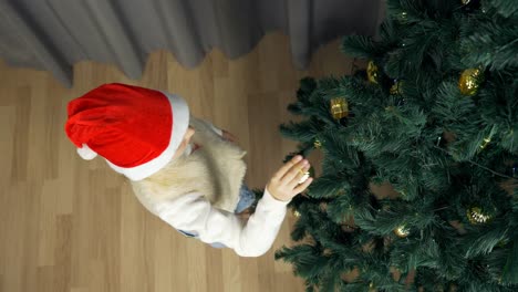 Slow-Motion-Draufsicht-erschossen.-Junges-Mädchen-hängt-goldene-Kugel-am-Weihnachtsbaum.-Mädchen-in-rote-Weihnachtsmütze-Christbaum-schmücken.