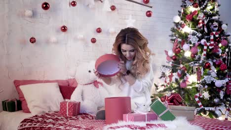 Young-attractive-woman-sits-on-a-bed-near-the-Christmas-tree-and-unpacks-presents