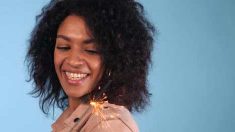 Sparkling-Bengal-fire-in-african-american-woman's-hand-on-blue-background.-Christmas-Holiday-Concept.-Young-pretty-girl-with-afro-hairstyle-celebrating,-smiling,-enjoying-time.-4k