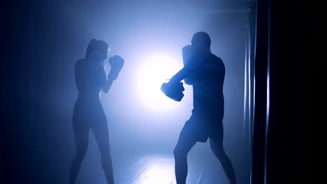 Silhouetten-von-eine-Boxerin-Stanzen-ein-Boxsack-mit-Boxhandschuhen-in-einem-verrauchten-Fitnessstudio