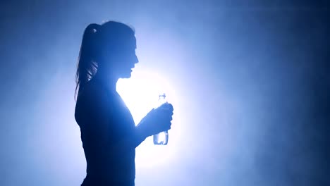 Boxeadora-sed-tomando-un-descanso,-bebiendo-de-la-botella-de-agua-después-del-entrenamiento-en-el-gimnasio-oscuro-con-humo.-Siluetas-de-una-mujer-del-boxeador-en-el-gimnasio-oscuro