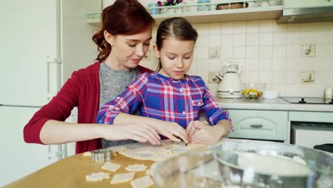 Fröhliche-Mutter-und-kleine-niedliche-Tochter-sprechen-und-Cookies-zusammen-mit-Bäckerei-Formen-Zuschneiden-Teig-während-der-Sitzung-in-der-modernen-Küche-zu-Hause-zu-machen.-Familie,-Essen-und-Leute-Konzept