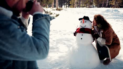 Couple-having-fun-taking-photos-with-snowman-in-the-snow