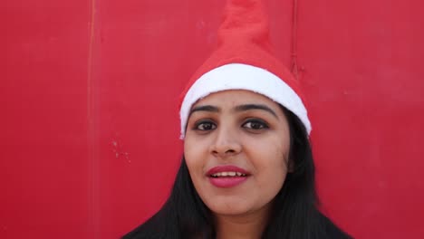 Young-Indian-woman-with-Santa's-hat-welcome-and-making-hand-gestures,-excited-and-super-happy,-with-a-matching-red-background,-handheld-gimbal-stabilized