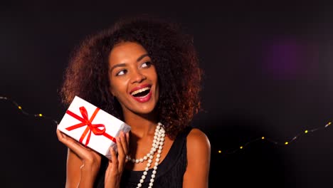 African-american-young-woman-in-party-dress-holding-gift-box-with-red-ribbon-and-bow-on-black-lights-background.-Girl-smiling,-she-happy-to-get-present-on-New-Year-or-Christmas