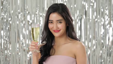 Beautiful-asian-woman-holding-glass-of-champagne-dancing-among-confetti-over-silver-glitter-background,-slow-motion