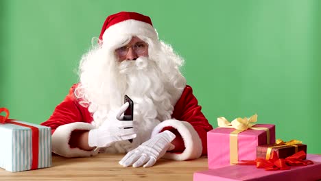 Smiling-Santa-Claus-showing-smartphone-in-camera,-gifts-on-the-table,-chromakey-in-the-background